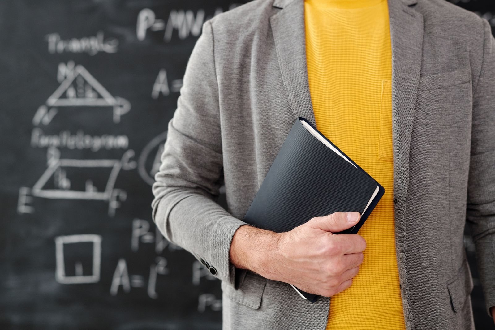 Docente a segurar um caderno junto de um quadro de sala de aula.