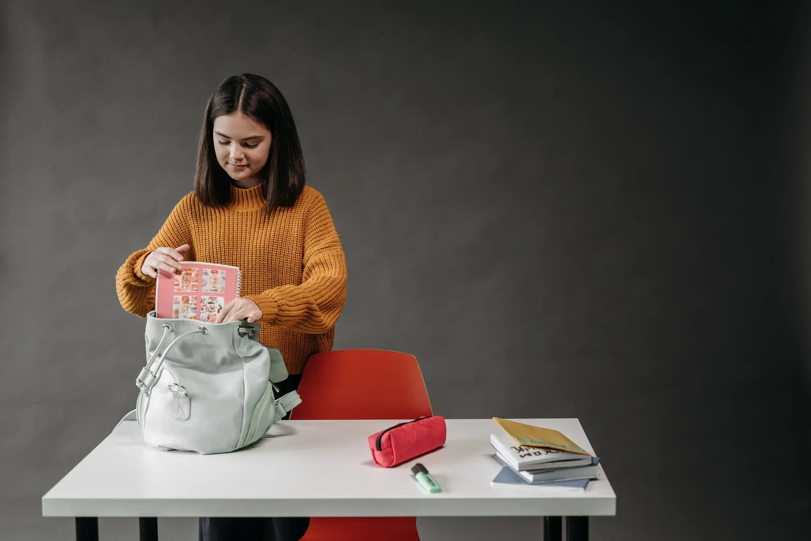 Uma estudante coloca um caderno na mochila, junto de livros e um estojo sobre a mesa.